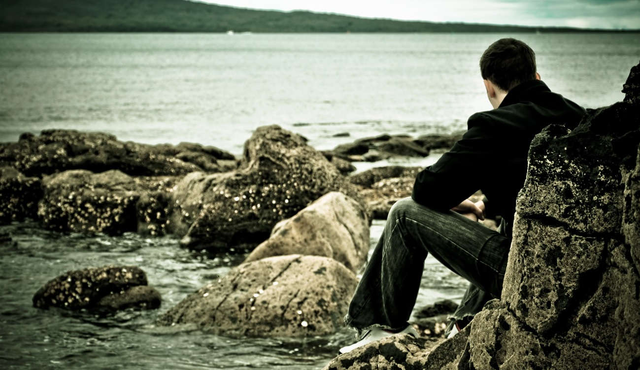 Hombre sentado de espaldas junto al mar en una playa rocosa en un día gris